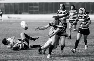 Black and white image of female rugby players on pitch.  As two lay on the grass pitch and five women running towards the rugby ball.