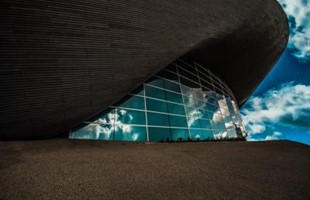 Image à l'extérieur du côté d'un bâtiment avec des panneaux de verre reflétant un ciel bleu et nuageux.