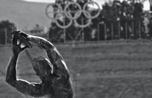 Black and white image of the back of a statue of an athlete, with sculpture of the olympic rings in the distance.